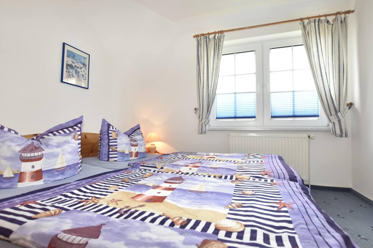 A bedroom with a nautical-themed bedspread, a framed picture on the wall, and a window with curtains.