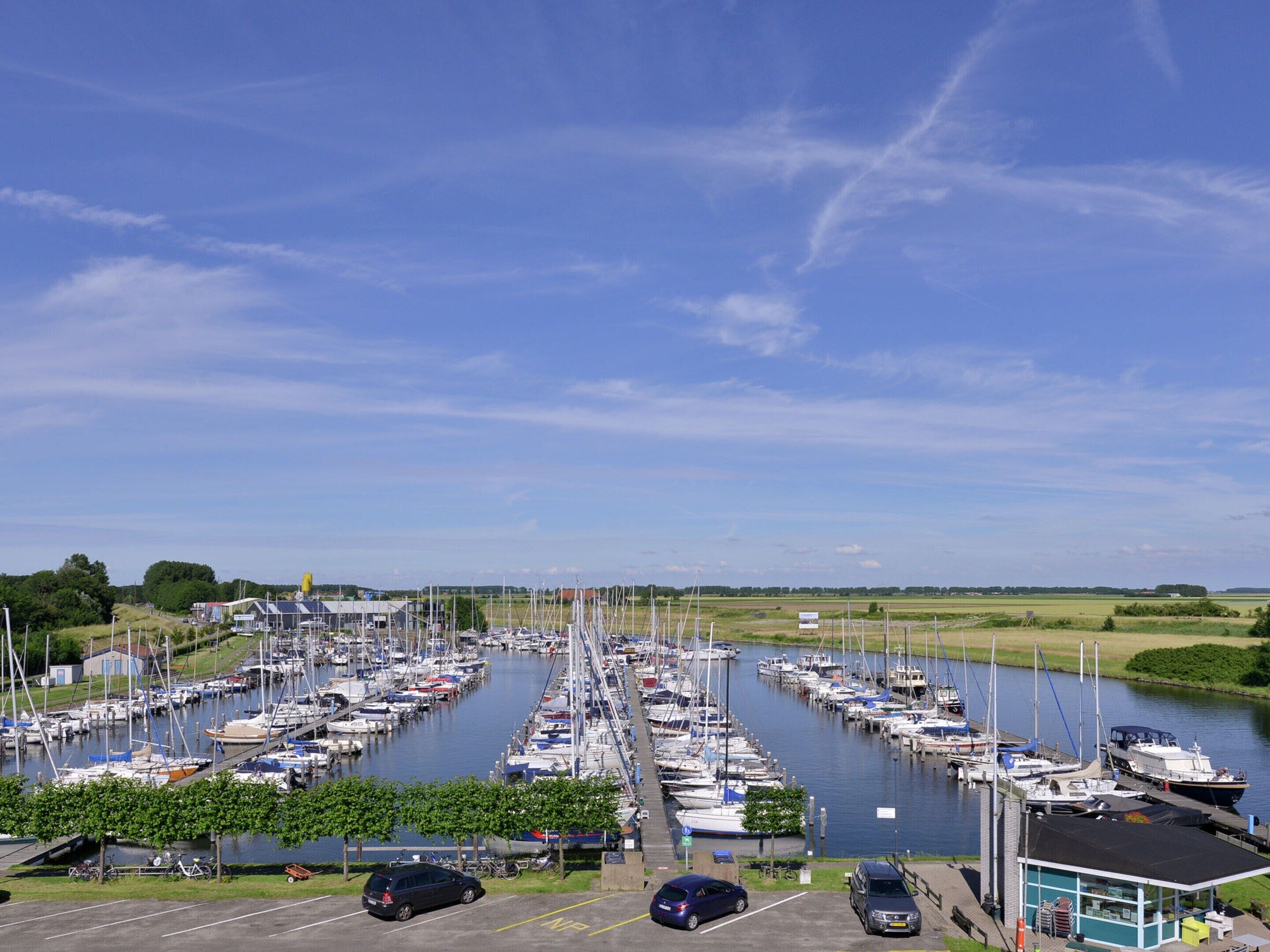 A marina with numerous boats docked in rows, a parking area with cars, and a clear sky.