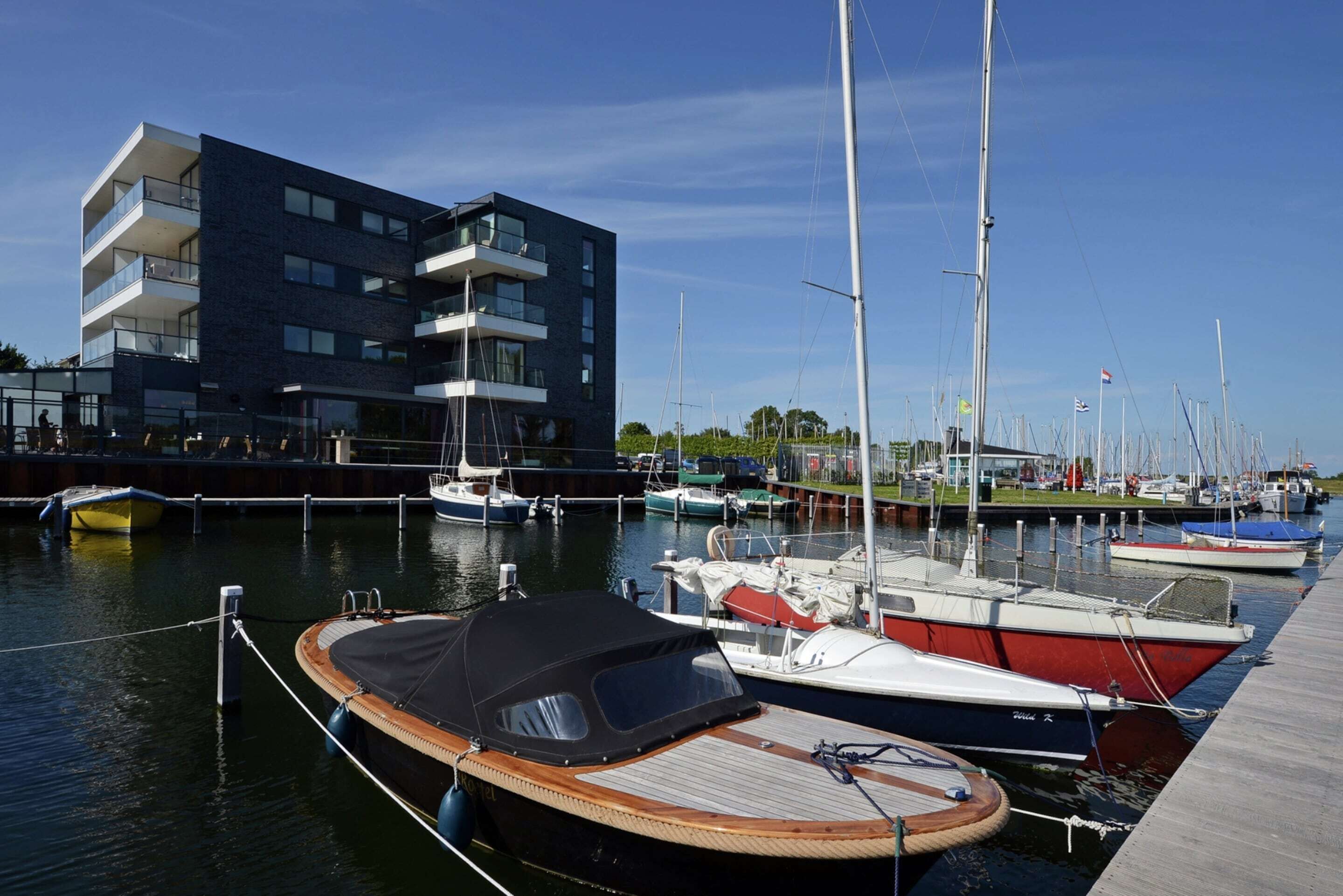 A marina with several boats docked, a modern building with balconies, and a clear blue sky.