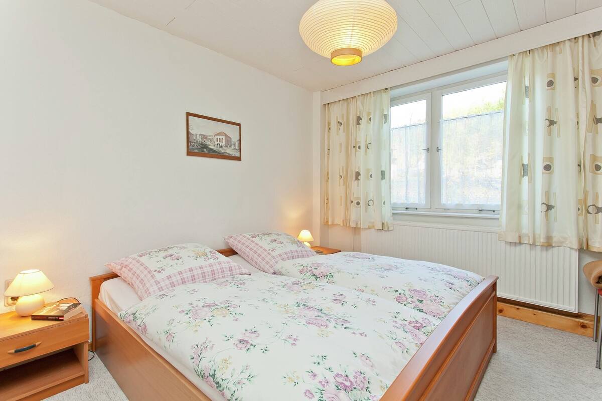 Ein Schlafzimmer mit einem hölzernen Bett, zwei Nachttischen mit Lampen, einem Fenster mit Vorhängen und einem gerahmten Bild an der Wand.