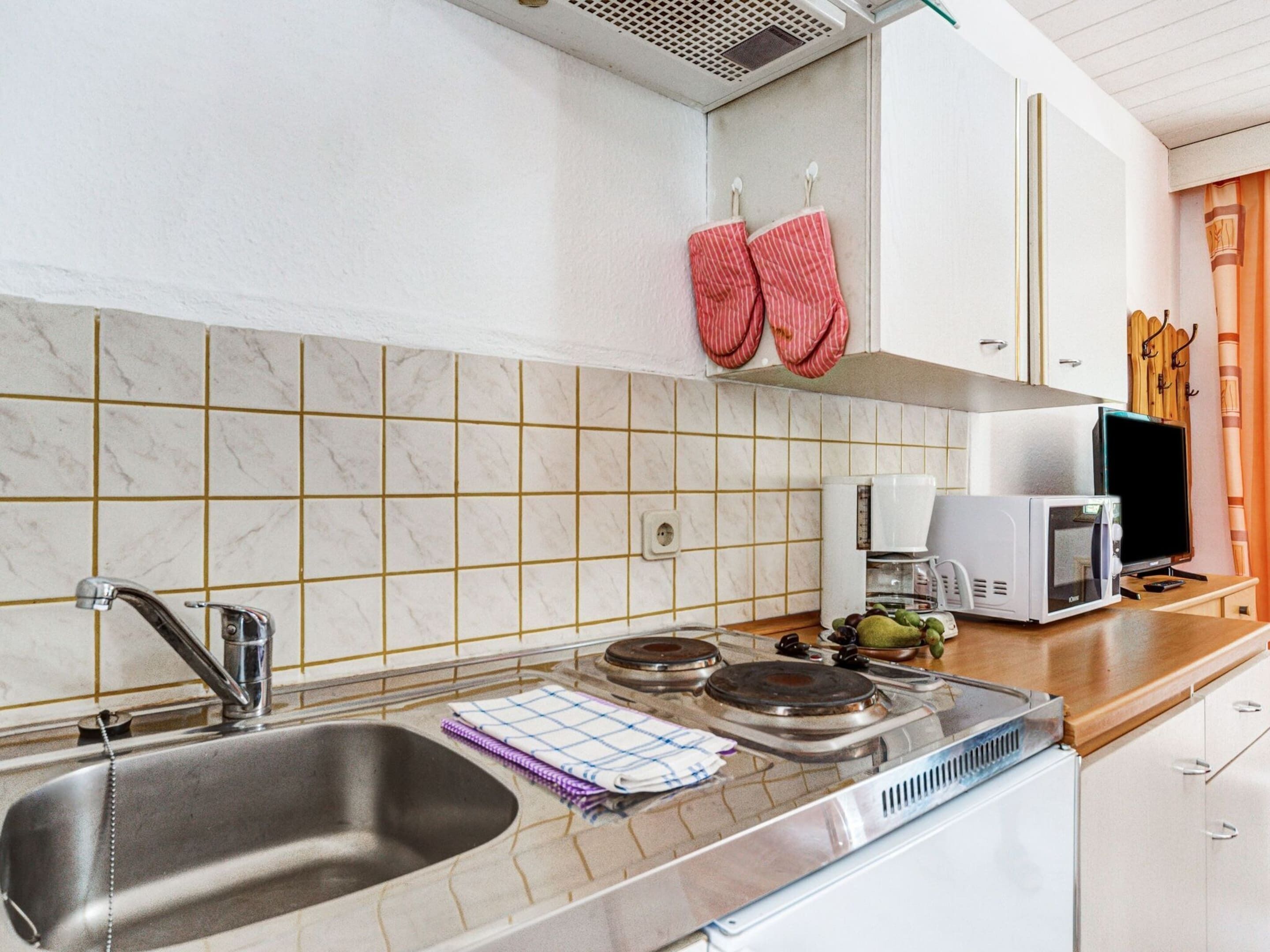 A kitchen with a sink, stove, and microwave. There are red oven mitts hanging on the wall.