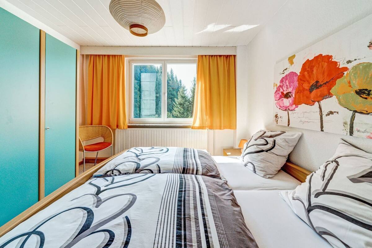Ein Schlafzimmer mit einem Bett, einem Sessel, einem Fenster mit Vorhängen und einer Wand mit einem Blumenbild.