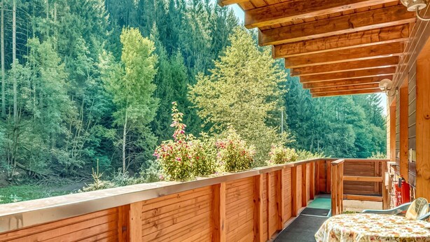 A wooden deck with a view of a forested area. There is a table with a floral tablecloth and two chairs. - Luxurious Apartment in Heubach Germany in the Forest (Masserberg)