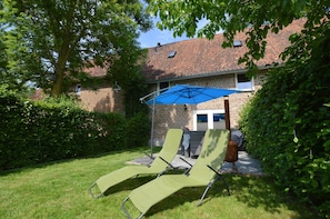 Un jardin avec une maison en briques, un parasol bleu et deux fauteuils de jardin verts.