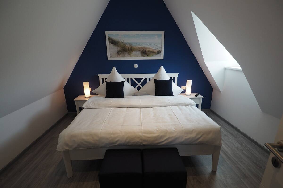 A bedroom with a white bed, two bedside tables with lamps, a painting on the wall, and a sloped ceiling.