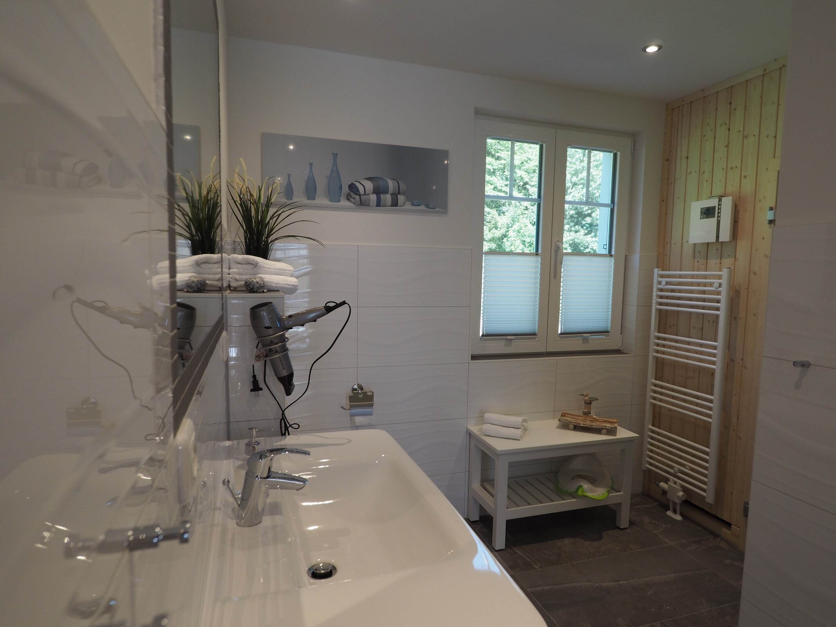 A modern bathroom with a large sink, a hairdryer mounted on the wall, a window with blinds, and a towel rack.