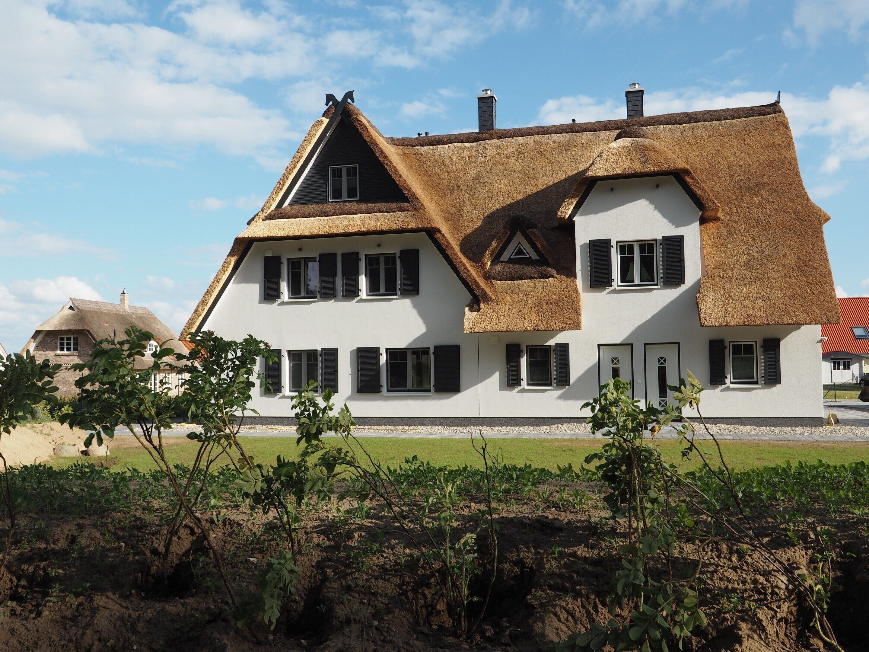 Uma casa grande e branca com telhado íngreme e várias chaminés, rodeada por um jardim bem cuidado e um campo ao fundo.