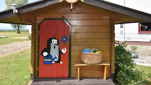 Eine Holzhütte mit roter Tür, einer bemalten Zeichentrickfigur, einem Schiffsrad und einem Korbkörbchen auf einer Bank.