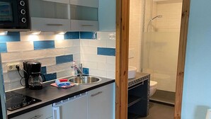 Eine moderne Küche mit Einbauofen, Kaffeemaschine und Spüle. Ein Holztürrahmen führt zu einem Badezimmer mit Dusche und Toilette.
