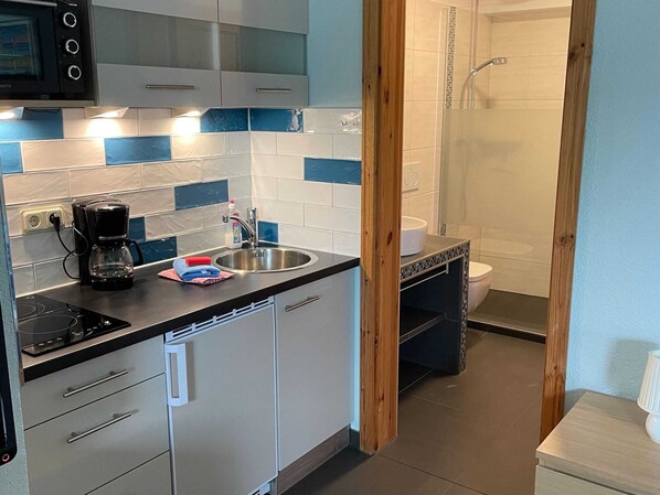 A modern kitchen with a built-in oven, a coffee maker, and a sink. There is a wooden-framed doorway leading to a bathroom with a shower and toilet. - Modern Apartment in Bastorf Near Sea (Bastorf)