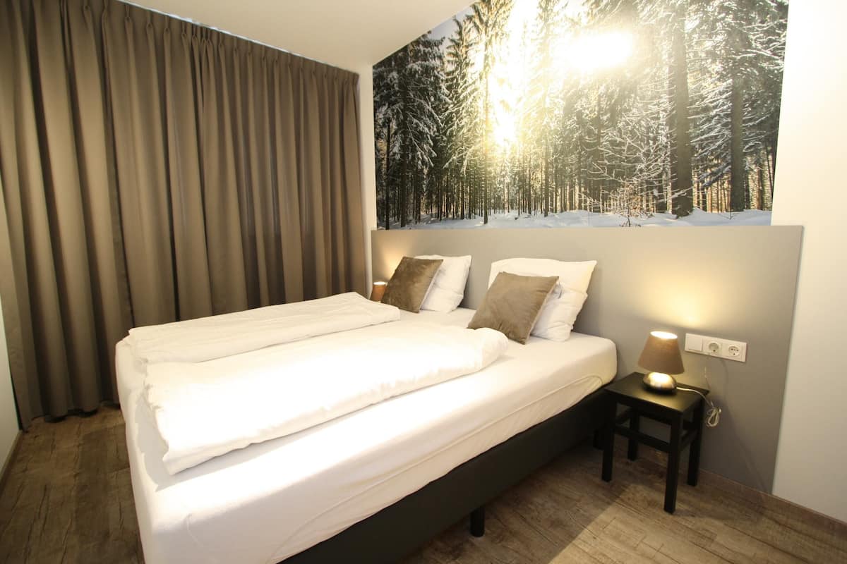 A neatly made bed with white linens and two pillows, a bedside table with a lamp, and a wall mural of a snowy forest scene.