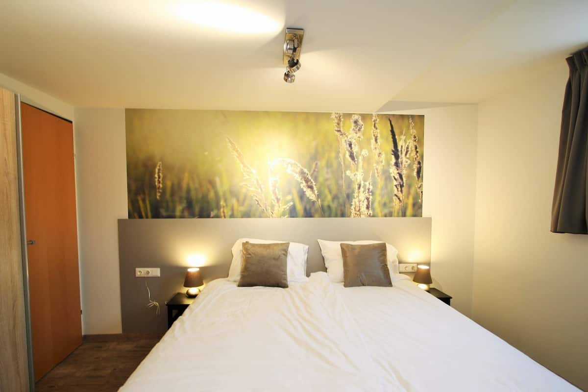 A large bed with white bedding and two bedside lamps, a wall-mounted artwork of tall grasses, and a wooden door.