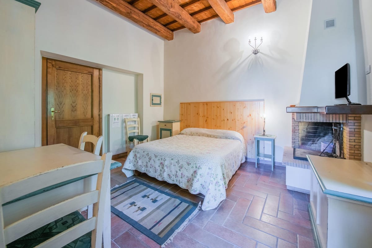 A bedroom with a wooden ceiling, a bed with a floral bedspread, a small table, a chair, a nightstand, a TV, and a fireplace.
