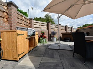 Eine Terrasse im Freien mit einem Holzgrill, einem großen Sonnenschirm und einer Sitzecke mit einem Korbstuhl.