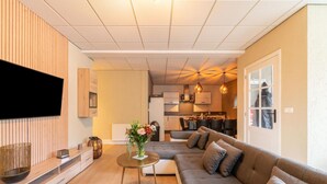 Ein modernes Wohnzimmer mit einer großen Couch, einem flachbildfernseher an der Wand, einem kleinen runden Tisch mit einer Blumenvase und einer Küche im Hintergrund.