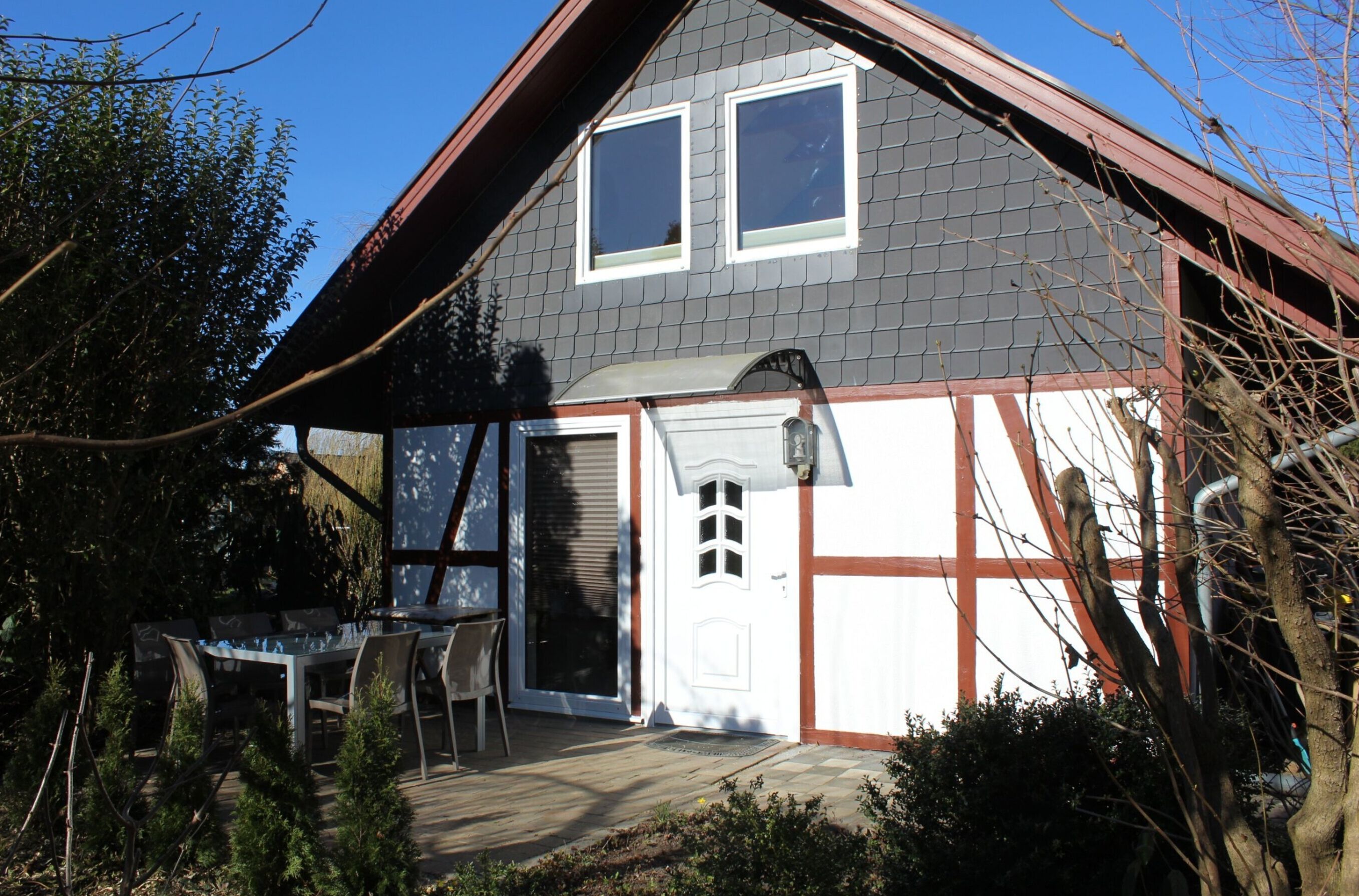 Een tweelaags huis met een grijs dak en witte details, met een voordeur, een klein terras met een tafel en stoelen, en een raam boven de deur.