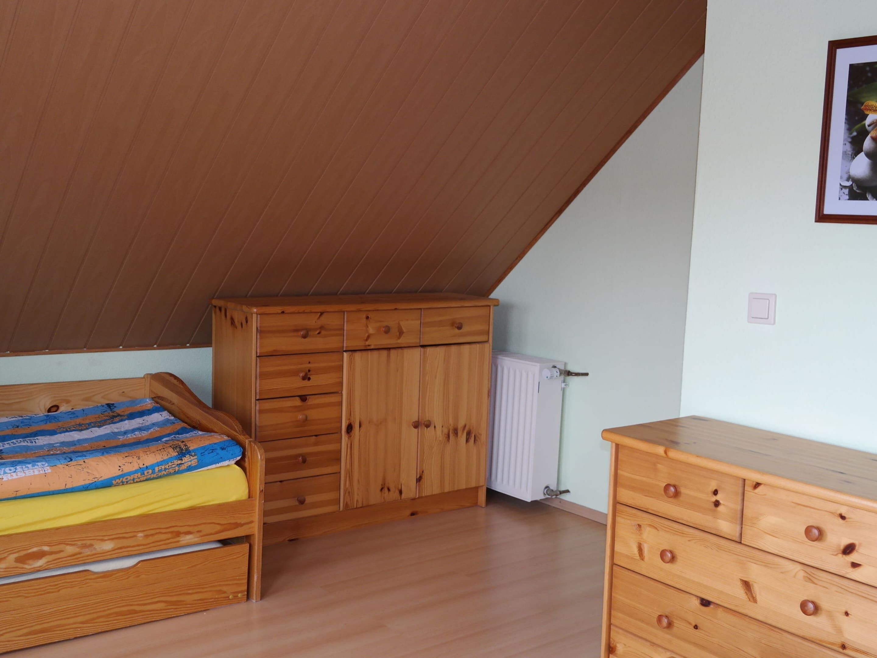 Une chambre avec un plafond en pente, un lit en bois, une commode en bois et un radiateur.