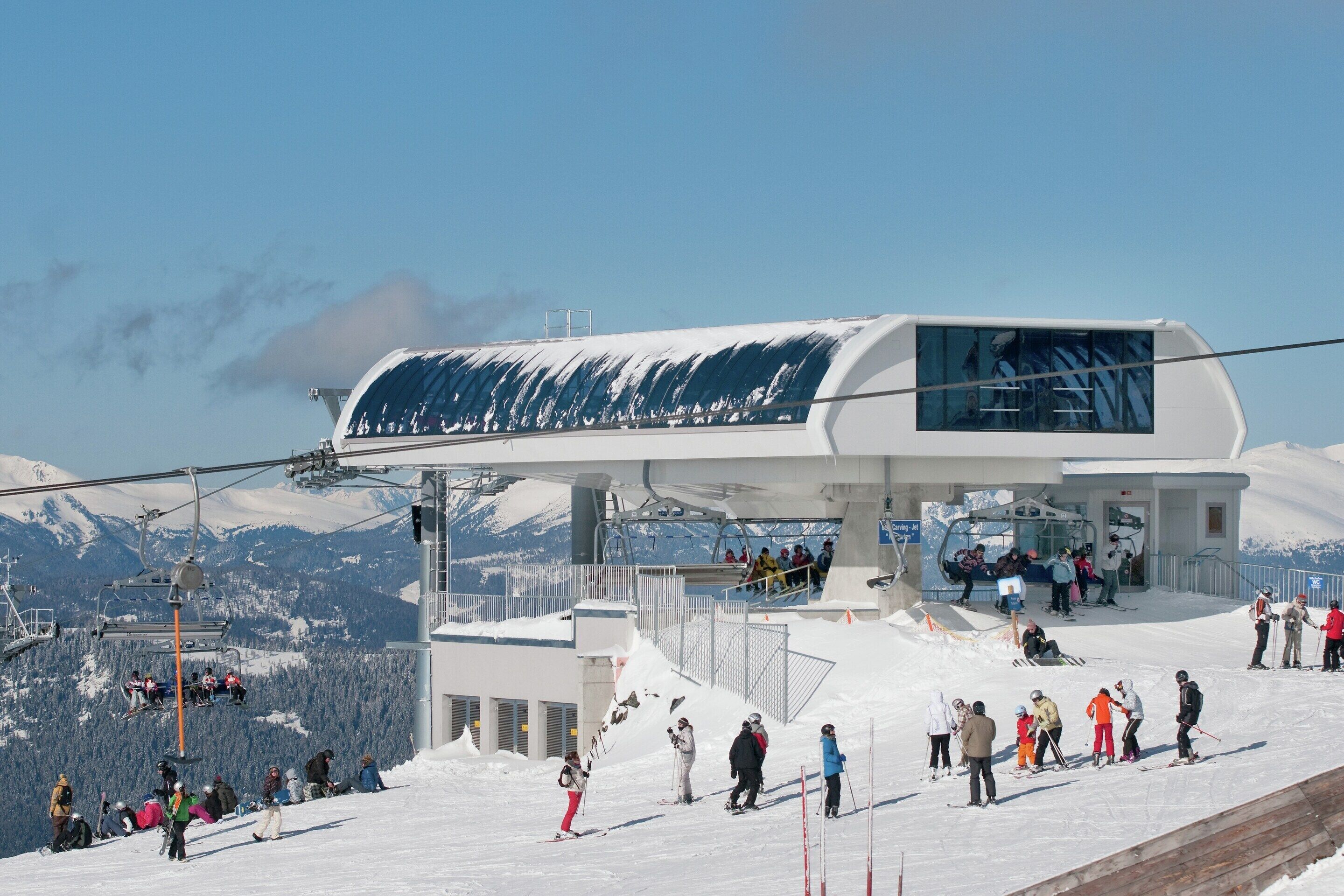 Un télésiège avec des personnes qui attendent, une zone abritée et des montagnes enneigées en arrière-plan.