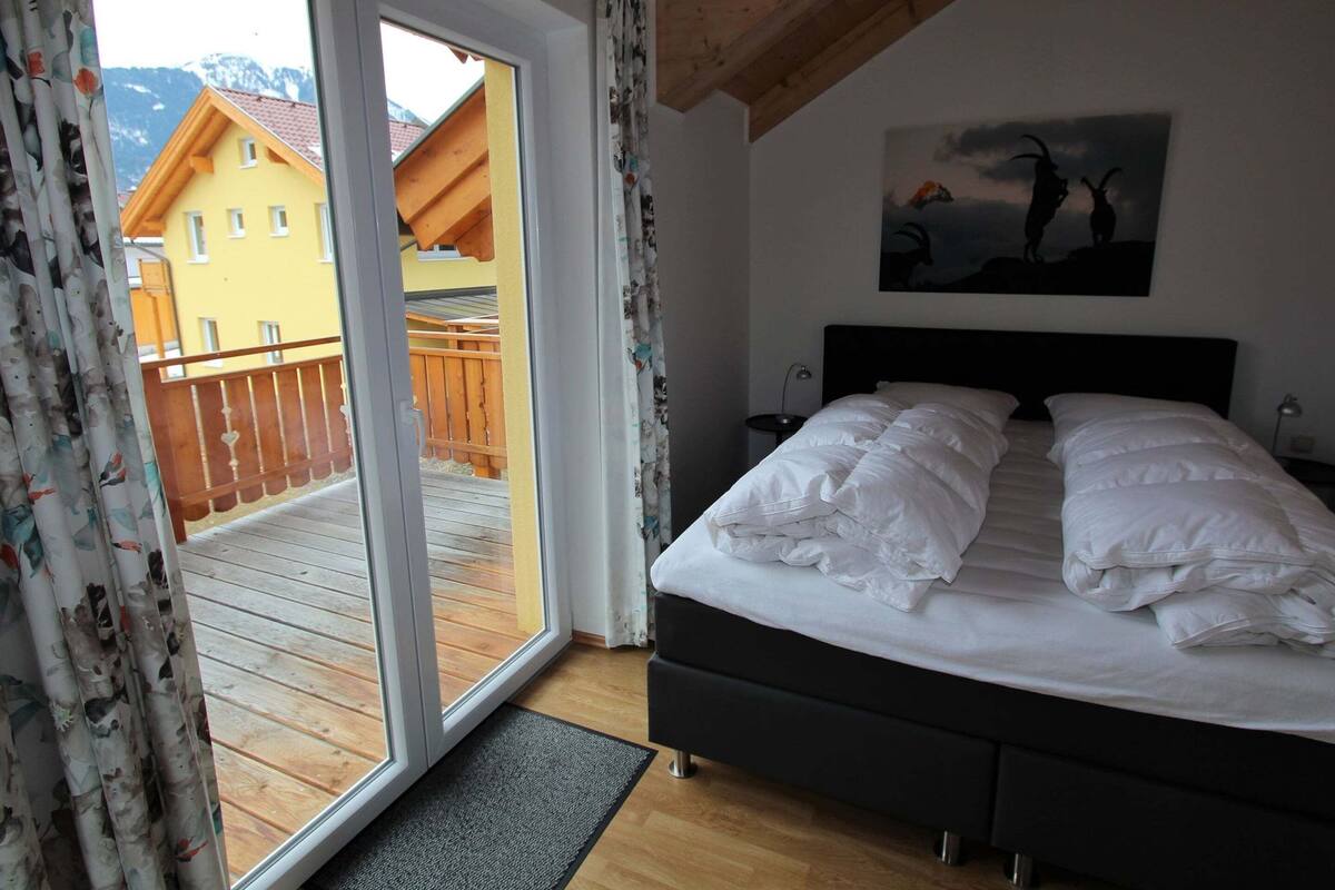 Ein Schlafzimmer mit Bett, Nachttisch, einem Gemälde und Blick auf eine Holzterrasse sowie Gebäude in den Bergen durch eine Schiebetür.