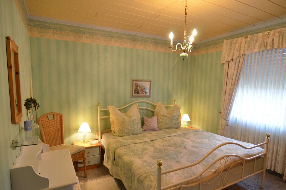 A bedroom with a bed, nightstand, chair, and a chandelier.