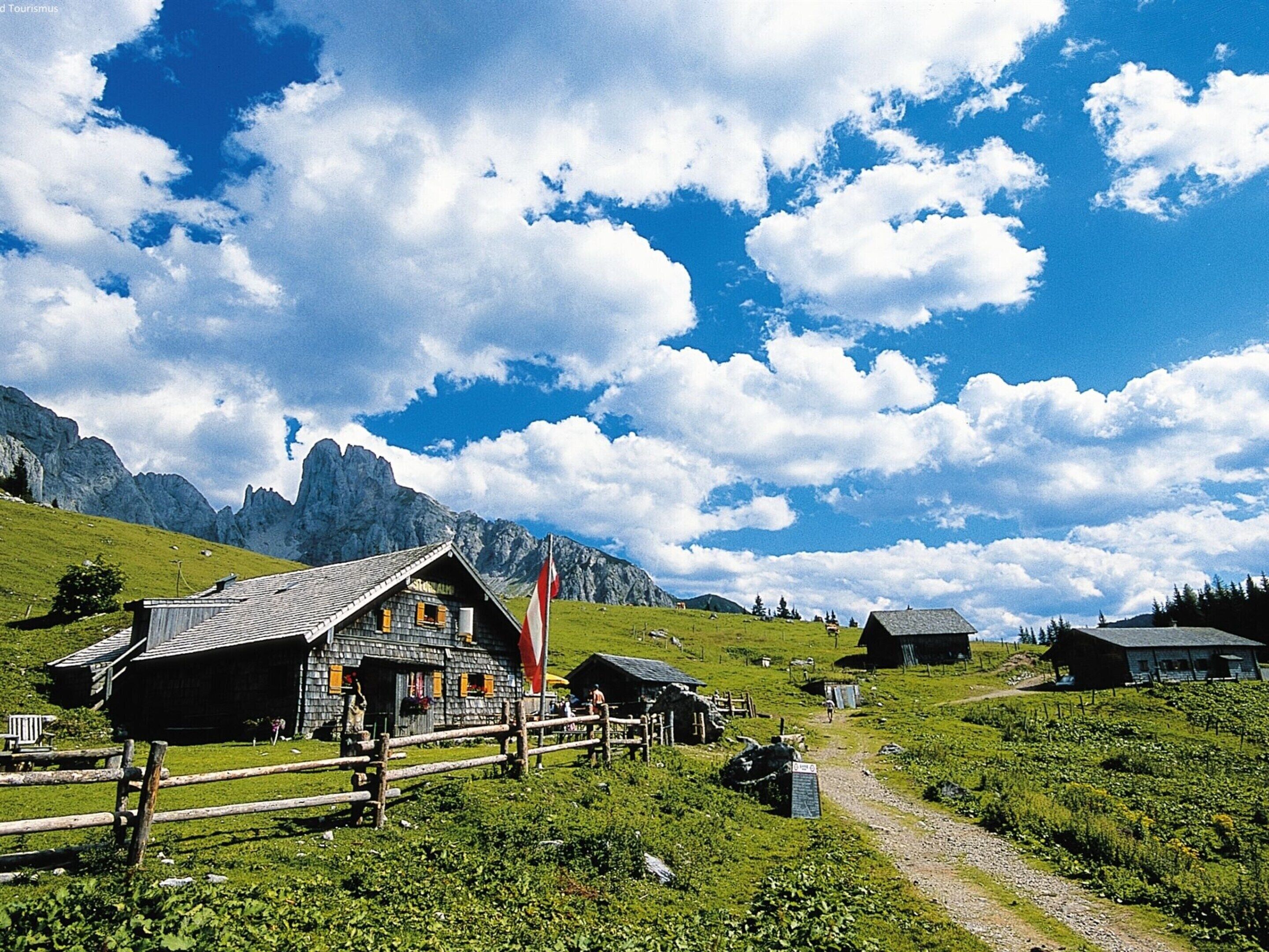 Un chalet traditionnel en bois, doté d’un toit à pente, entouré d’une clôture en bois, et se dressant sur un fond de montagnes et d’un ciel dégagé.