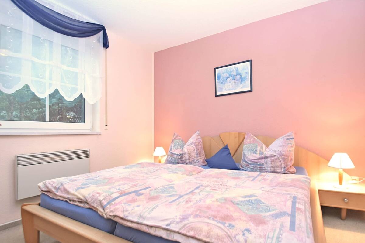 Ein Schlafzimmer mit einem Bett, zwei Nachttischen, einem Fenster mit Vorhang, einem Heizkörper und einem gerahmten Bild an der Wand.