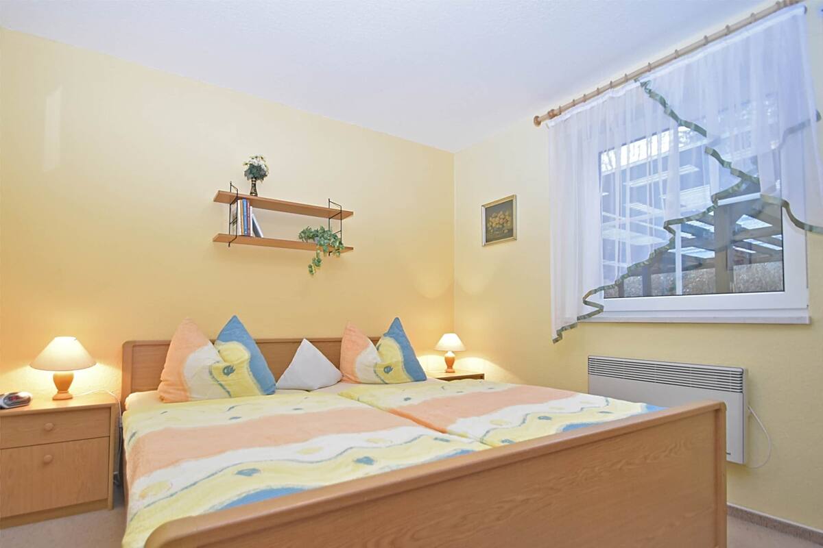 Ein Schlafzimmer mit Bett, Nachttischlampen, einem Fenster mit Vorhängen, einem Heizkörper, einem Regal mit Büchern und einer Pflanze sowie einem Bild an der Wand.