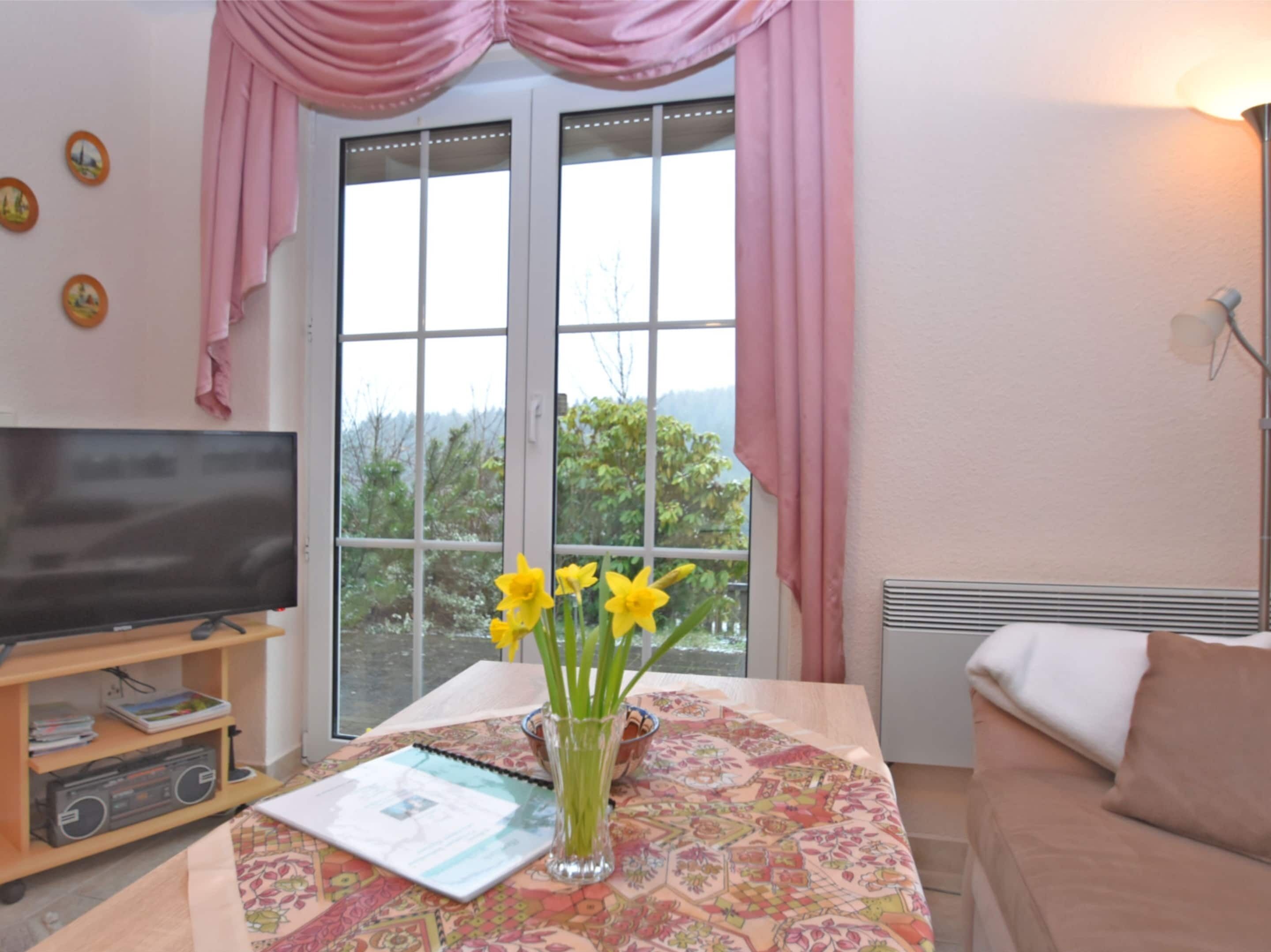 A living room with a television, a table with a vase of flowers, and a view of greenery outside.