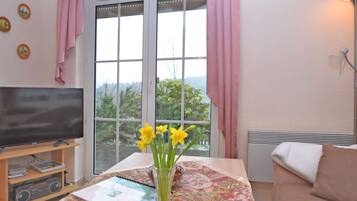 A living room with a television, a table with a vase of flowers, and a view of greenery outside.