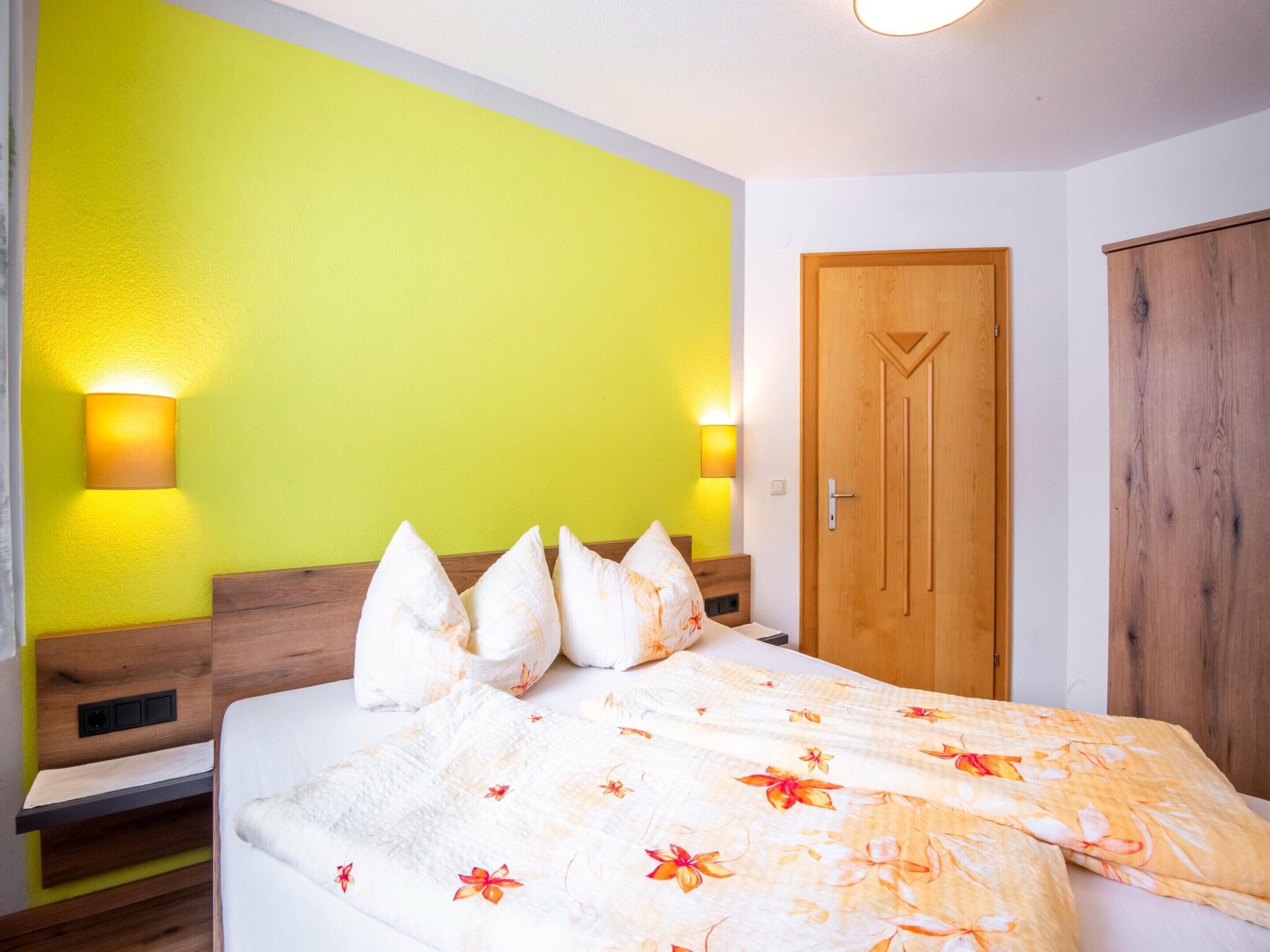 A hotel room with a bed, a wooden wardrobe, a wooden door, and two wall-mounted lights.