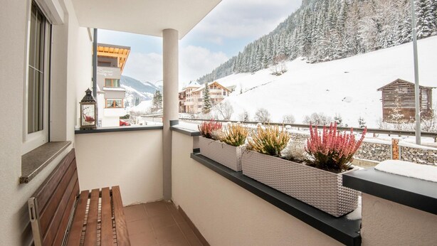Un balcón con banco de madera, plantas en macetas y vista a un paisaje montañoso nevado.