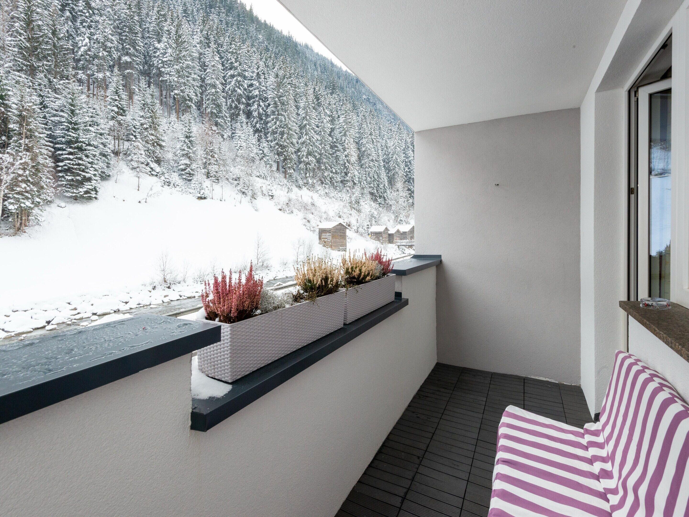 Un balcon avec vue sur des arbres couverts de neige et une rivière. On y trouve un pot de fleurs blanc avec des plantes et un coussin à rayures.