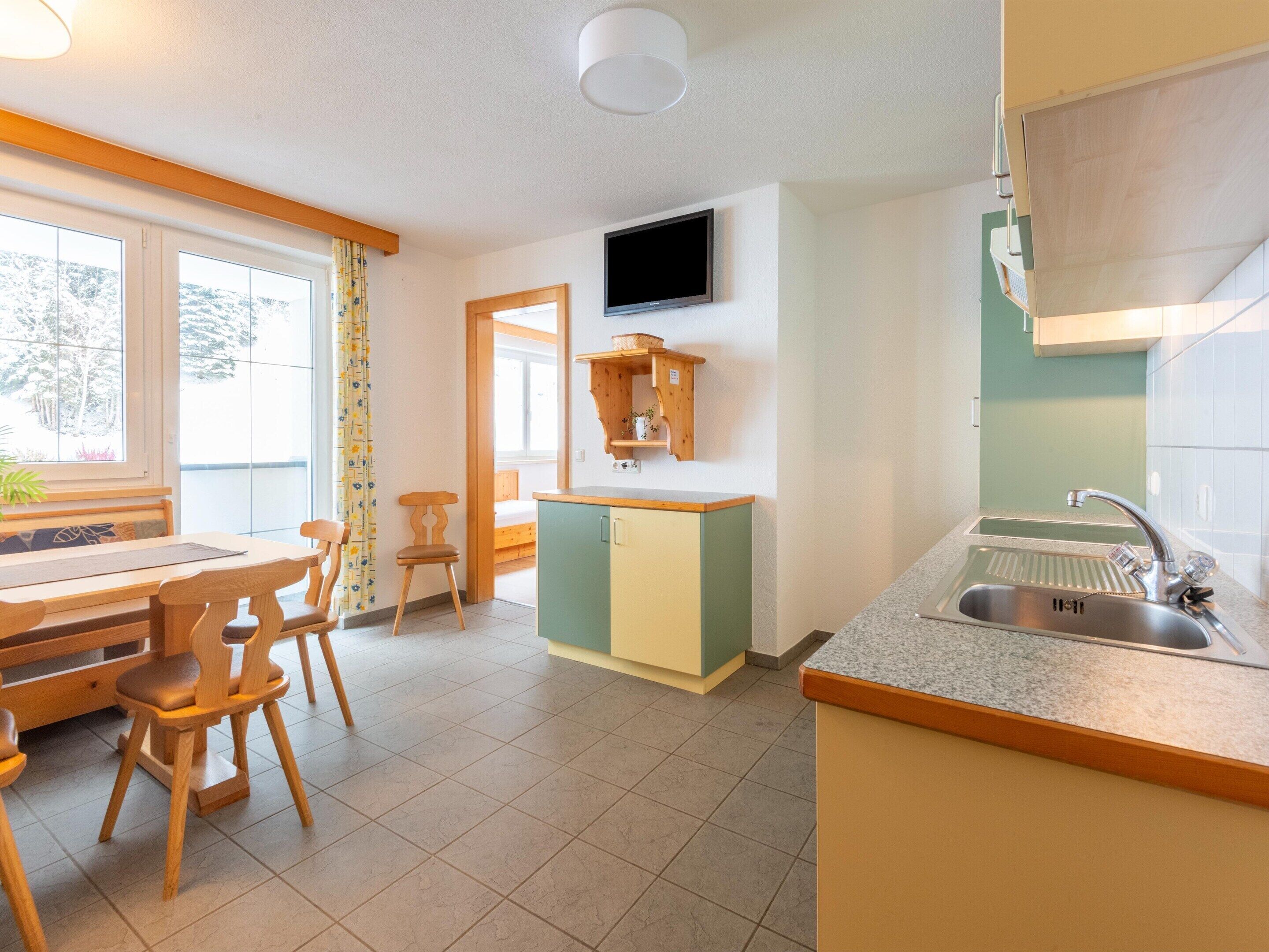 A compact kitchen with a dining area, a TV mounted above the counter, and a window with curtains.