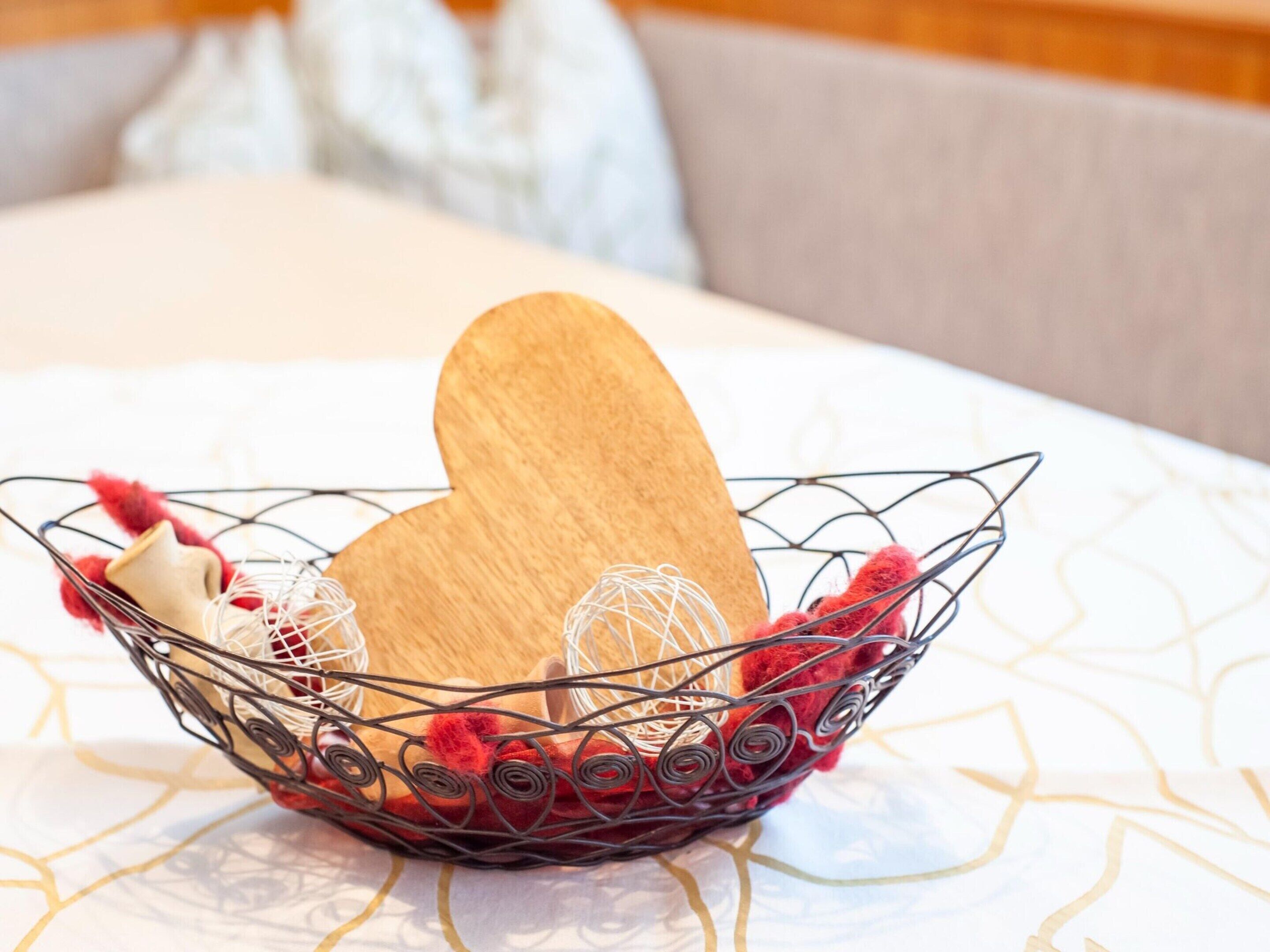 A metal wire basket containing a wooden heart-shaped object, a red fluffy toy, and a white spherical object on a patterned tablecloth.