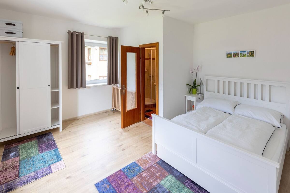 Une chambre avec un lit blanc, une armoire, une salle de bain avec douche et un tapis coloré.