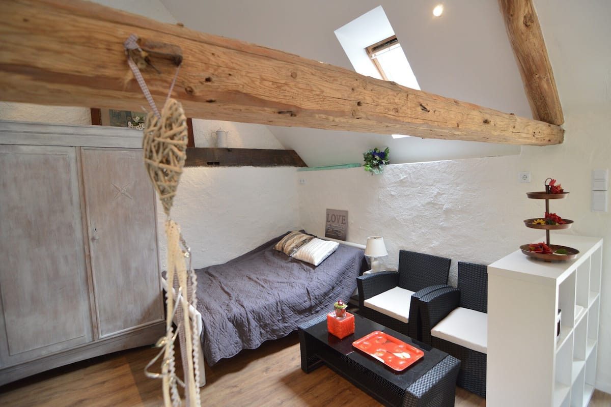 A compact room with a bed, a small table, a chair, a shelf with decorative items, and a wooden ceiling beam.