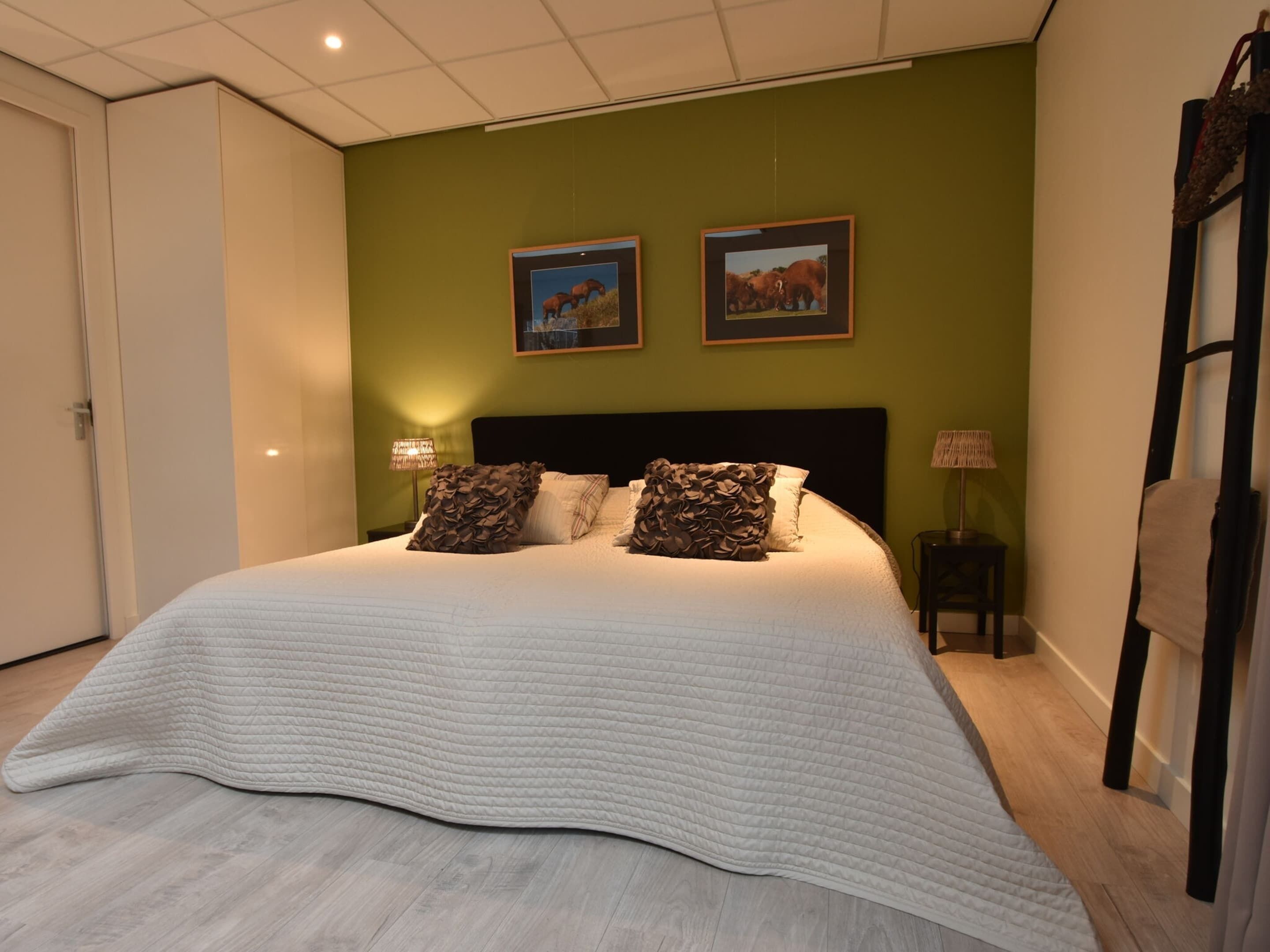 A bedroom with a large bed, two wall-mounted pictures, bedside lamps, and a wooden floor.