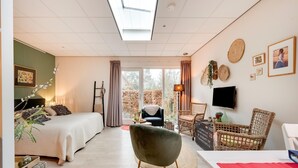Une pièce spacieuse comprenant un lit, un fauteuil vert, une table en osier et un téléviseur fixé au mur.