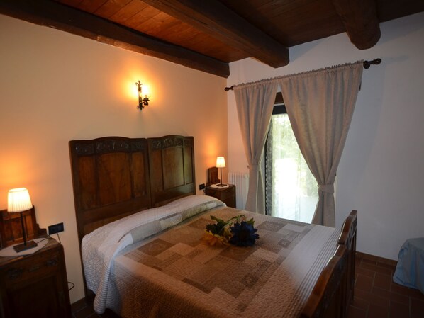 A bedroom with a wooden headboard, a bedside table with a lamp, a window with curtains, and a floral arrangement on the bed. - Cozy Holiday Home in Modigliana Italy With Garden (Modigliana)