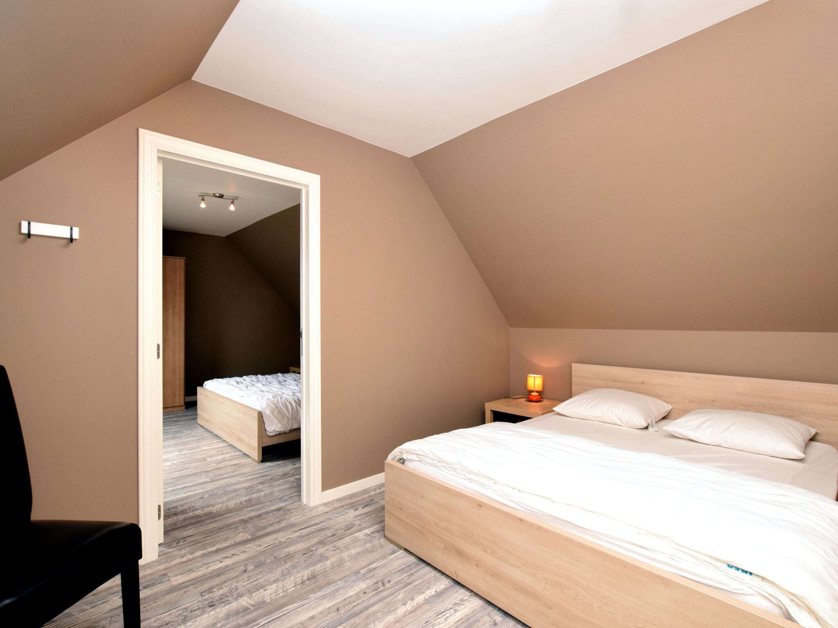A bedroom with a sloped ceiling, a bed with white bedding, a nightstand with a lamp, and a chair.