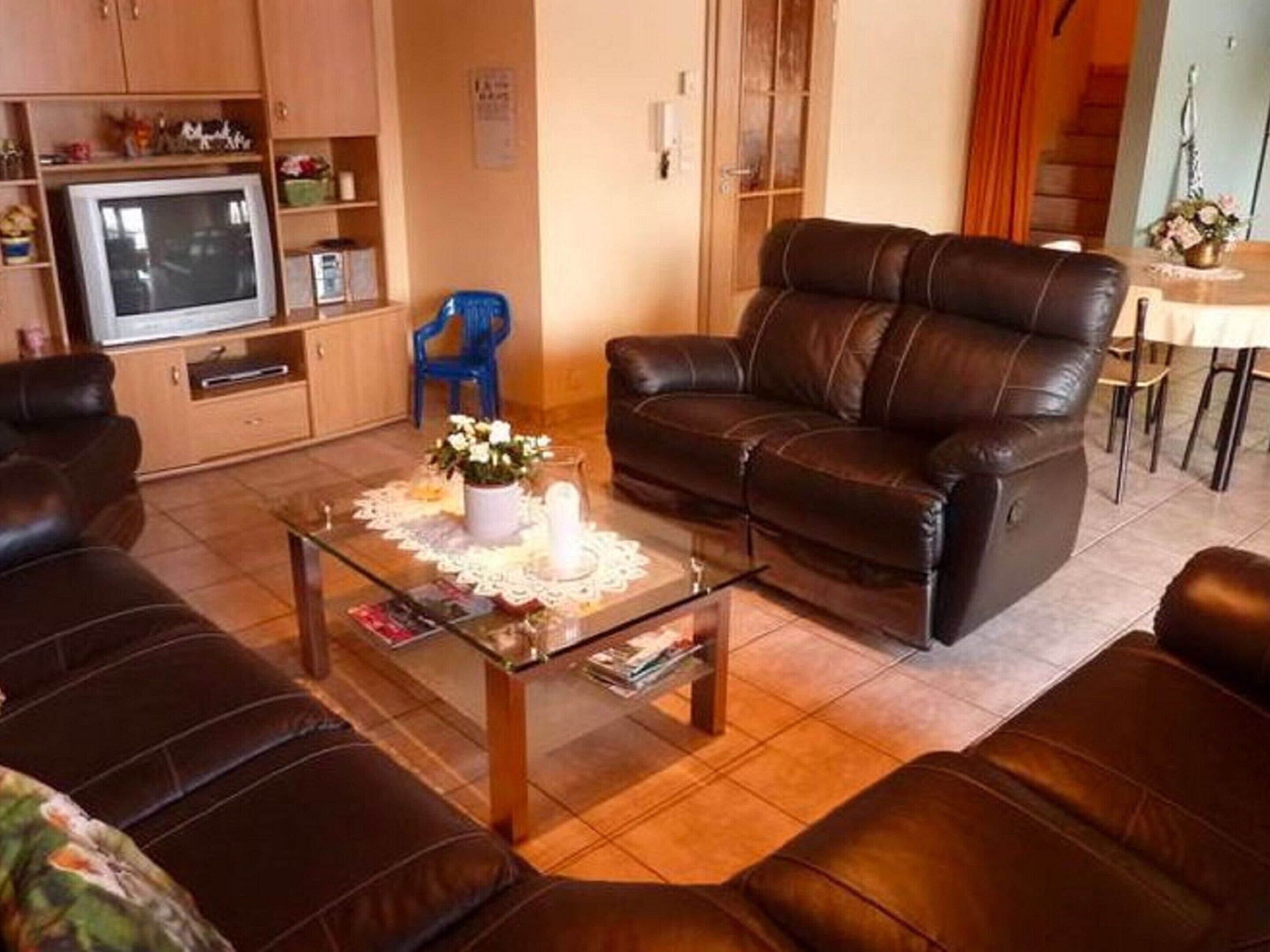 A living room with a brown leather sofa, a glass coffee table with a white doily, a blue chair, and a television on a wooden cabinet.
