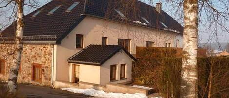 A two-story house with a stone facade and a tiled roof, surrounded by bare trees and a snow-covered ground.