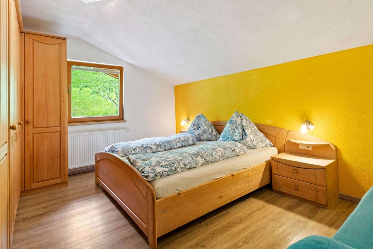 Un dormitorio con una cama de madera, una ventana con vistas al follaje, una pared amarilla y un armario de madera.