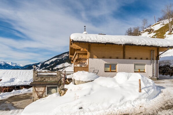 Un chalet de madera con techo nevado, un porche pequeño y una vista de montañas nevadas al fondo.