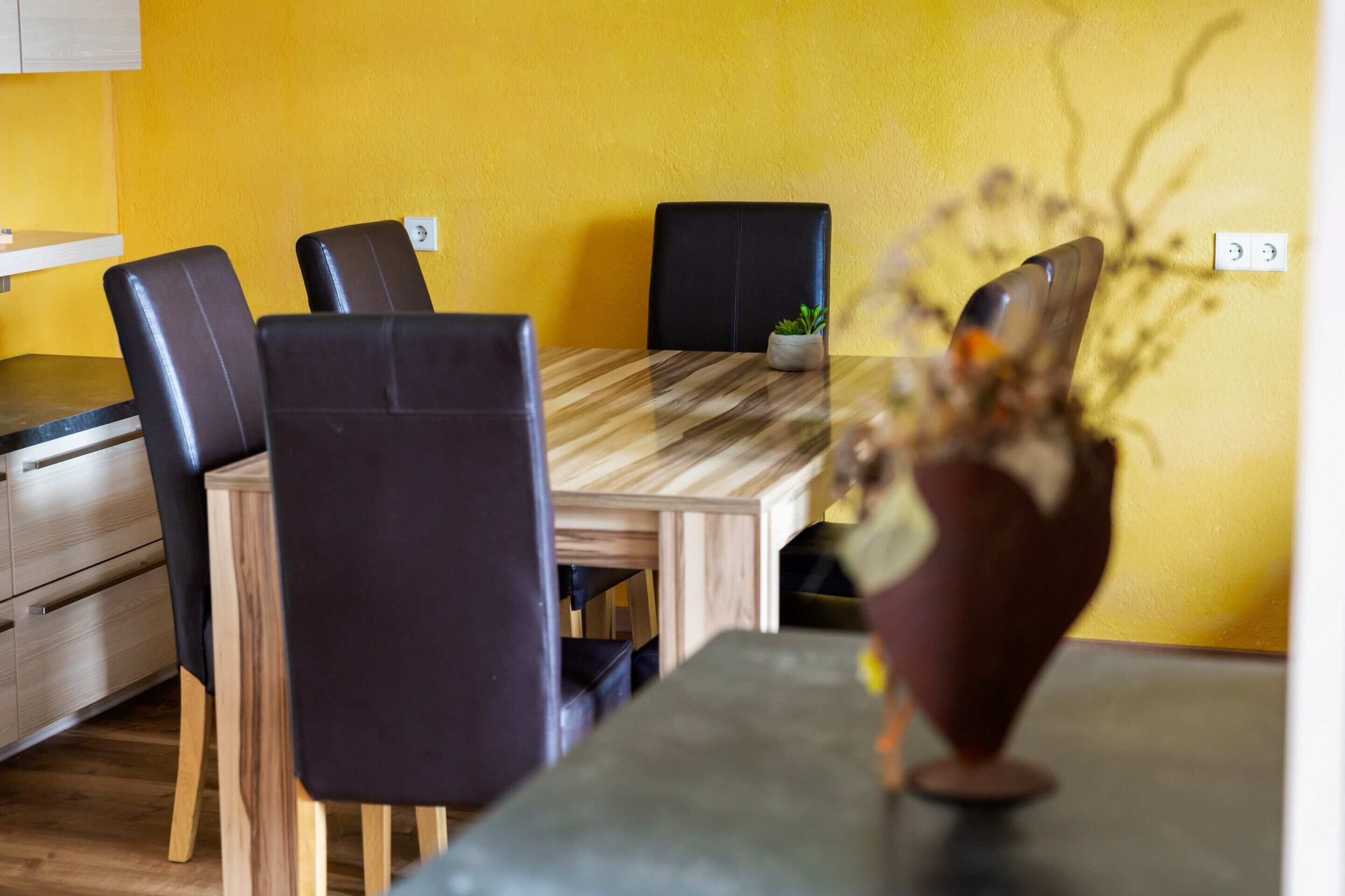 Un comedor con una mesa de madera y sillas negras, una planta pequeña en maceta y un jarrón con flores.