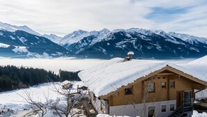 A chalet with a snow-covered roof, a wooden exterior, and a view of snow-capped mountains in the background. - Sunlit Apartment Near Ski Area in Hollersbach im Pinzgau (Hollersbach im Pinzgau)