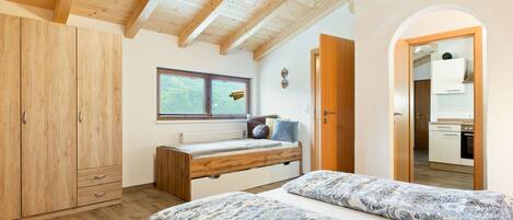 Une chambre avec un lit en mezzanine, des poutres apparentes en bois au plafond, une fenêtre, un bureau intégré et un coin cuisine.