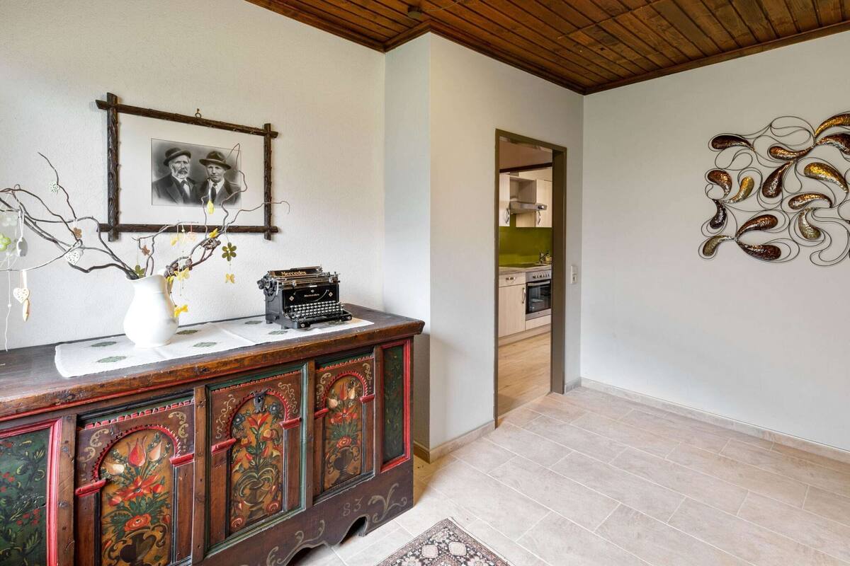 Une pièce avec un plafond en bois, une machine à écrire ancienne posée sur un meuble en bois, une photographie encadrée accrochée au mur et une cuisine visible à travers une porte ouverte.