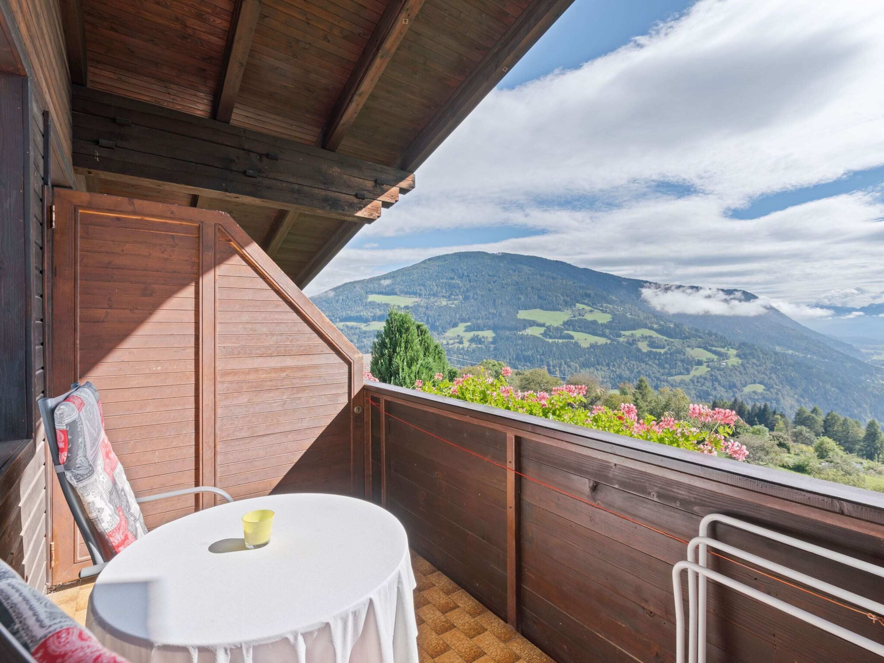 A wooden balcony with a table and chairs, overlooking a mountainous landscape with greenery and flowers.