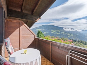 A wooden balcony with a table and chairs, overlooking a mountainous landscape with greenery and flowers.